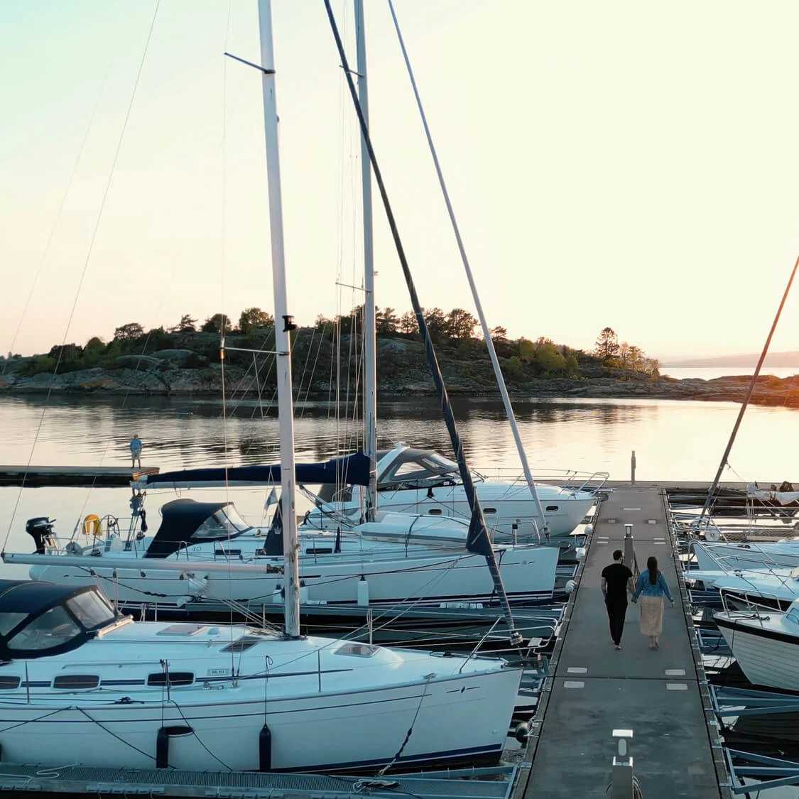 Båter fortøyd ved en marina med to personer som går på bryggen mens solen går ned i bakgrunnen, omgitt av rolig vann og steinete kystlinje.