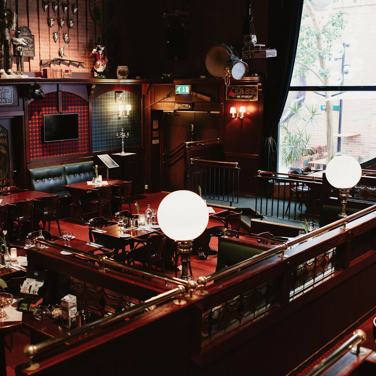Restaurant interior features vintage decor, wooden furniture, and dim lighting. Signs read: "British Rail North London" and "Beware of Trains." A large window shows outdoor plants. Tables are set with flowers and candles.
