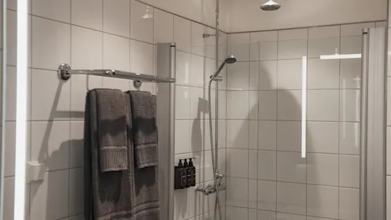 Ett rent och modernt duschutrymme med vita kaklade väggar, ett regnduschhuvud, en handdusch och fräscha handdukar på en ställning.