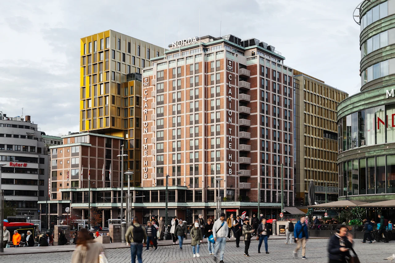 Folk går på en bygade foran en høj bygning med teksten CLARION THE HUB, omgivet af andre bystrukturer.