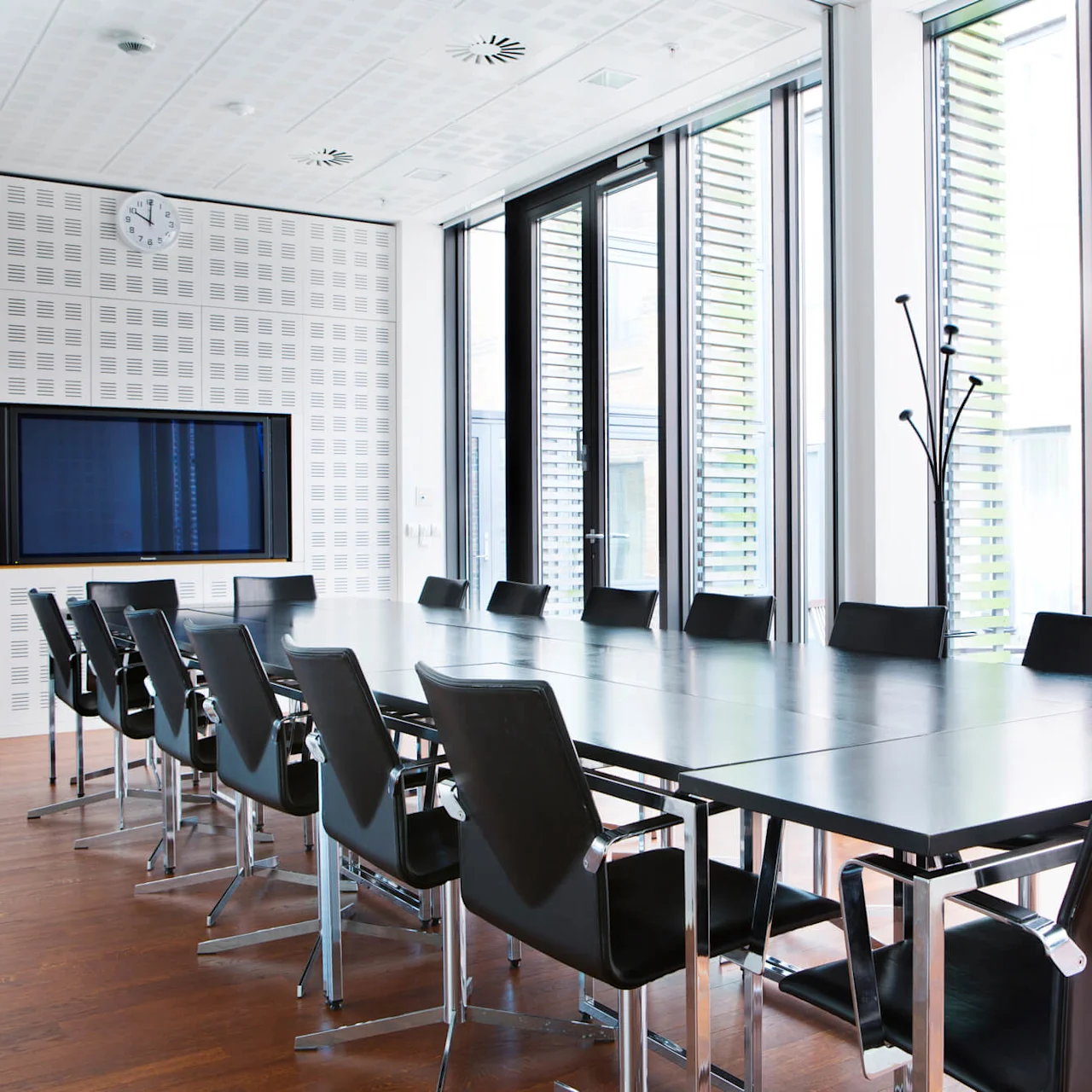 A modern meeting room with a long black table surrounded by black chairs with chrome details. The room features large windows allowing natural light and a wall-mounted TV.