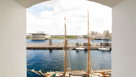 Utsikt genom en valvbåge över en hamn med en röd segelbåt förtöjd, moderna byggnader och andra båtar i bakgrunden.