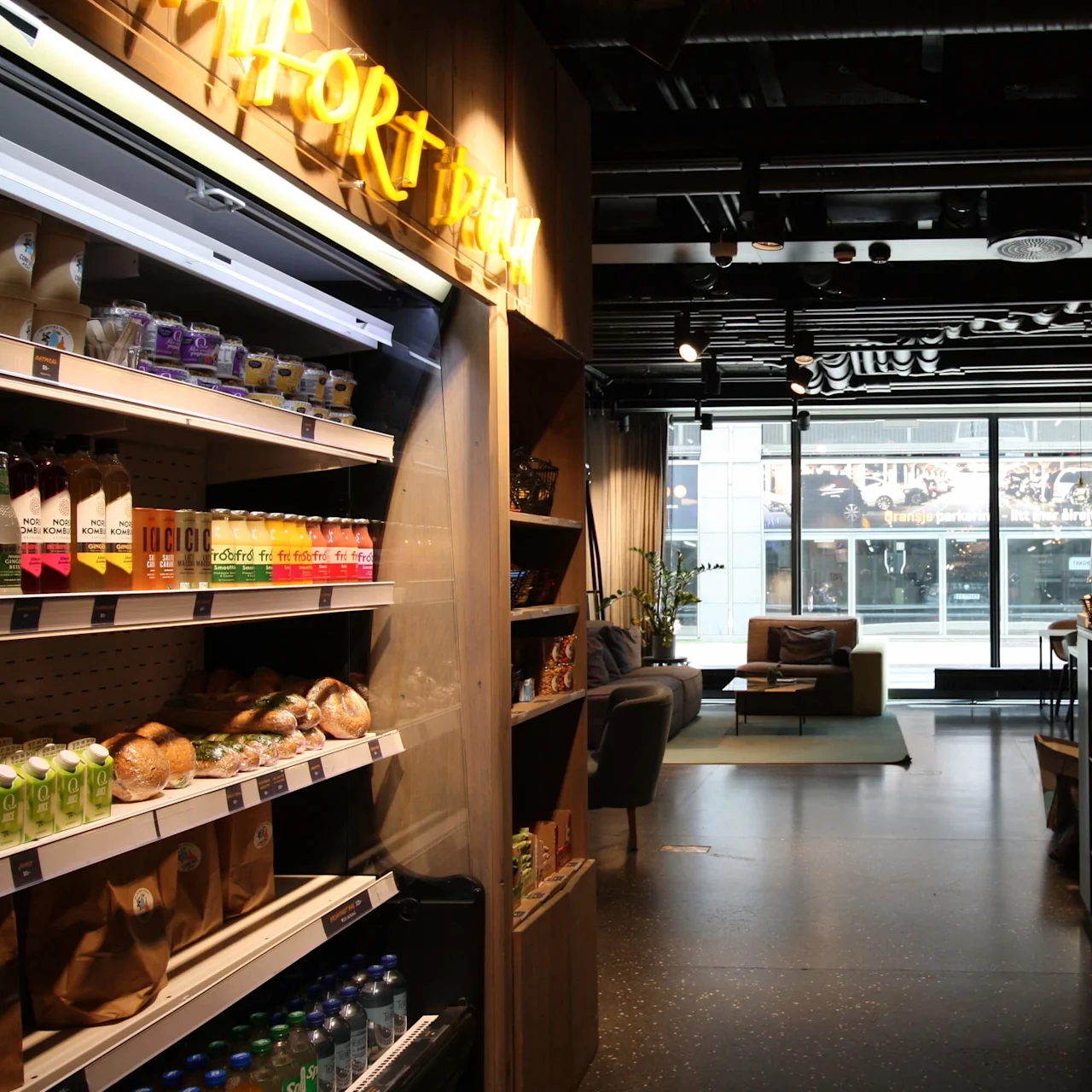 Shelves display drinks, snacks, and baked goods beside a bar with hanging lights and a cash register; a cozy seating area with sofas is visible through large windows. Text: "GRAB & GO." 