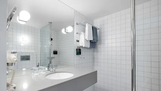 Et moderne bad med en hvit benkeplate under et stort speil, med en vask, glasskopper, en hårføner og håndklær. Sterkt lys reflekterer fra flislagte vegger.