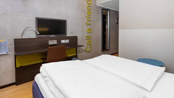 A bed with white linens faces a wall-mounted TV above a desk in a minimalist hotel room. Text on the wall reads "Call a friend".