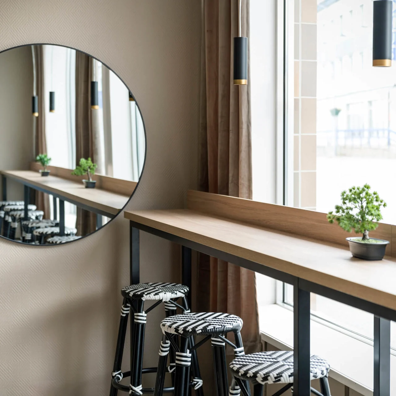 Round mirror reflecting a row of patterned stools beneath a wooden counter holding a small potted plant, opposite a large window. Modern pendant lights hang from the ceiling.