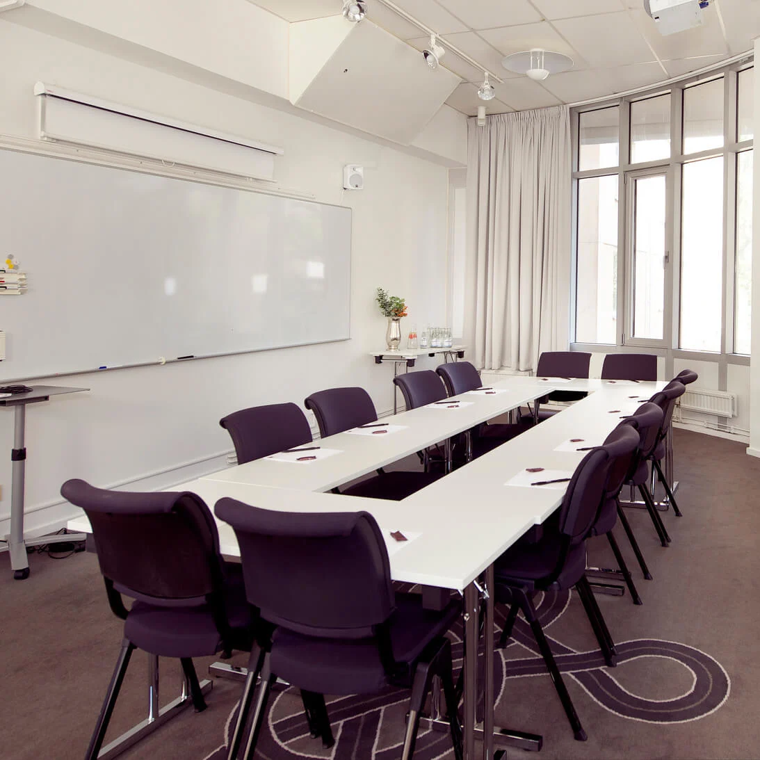 A conference room with a long table and chairs, bright from window light, with a whiteboard and a luxurious armchair.