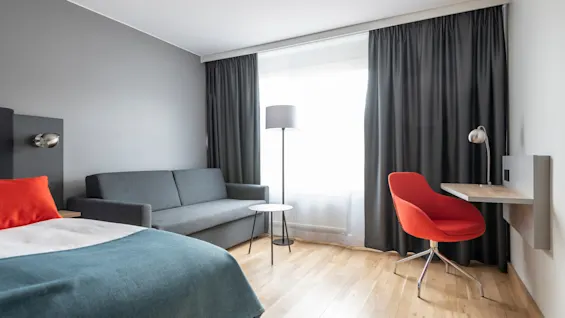 En grå soffa står i ett modernt rum med en golvlampa och litet bord vid fönstret. En orange stol är placerad vid ett skrivbord.