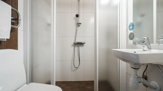 Dusch med krombeslag står i ett kaklat badrum, omgiven av glasväggar. En vit handfat och toalett finns i närheten. Klistermärken på glaset lyder "GO EASY ON THE PLANET. CONSUME LESS WATER".