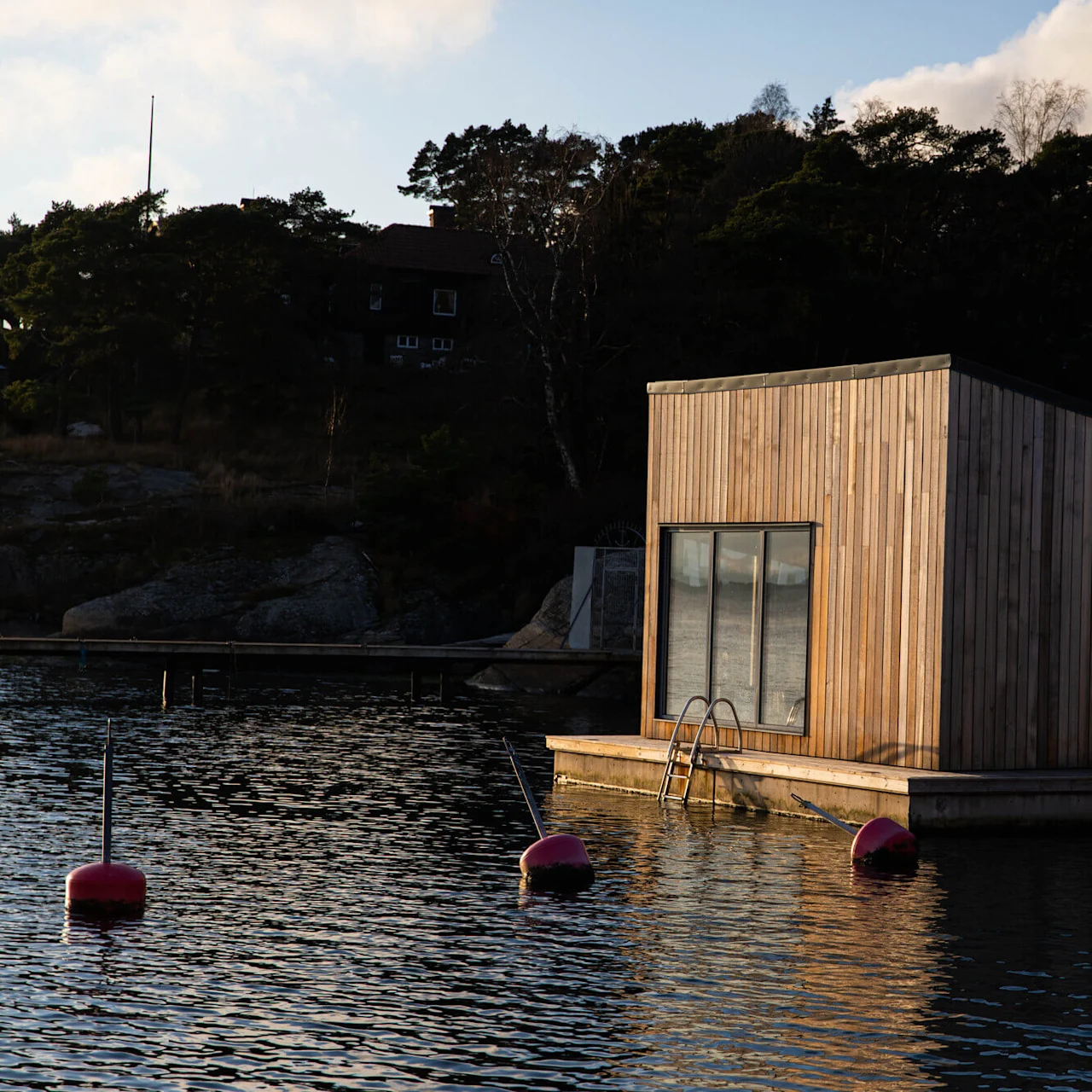 A modern wooden floating building with large windows is anchored on calm water, surrounded by trees and houses in the background. Pink buoys float nearby; two flags are visible.