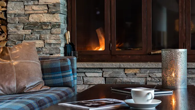 A cup on a wooden coffee table, near a lit candle and magazines, with a plaid sofa and stone fireplace in a cozy living room.