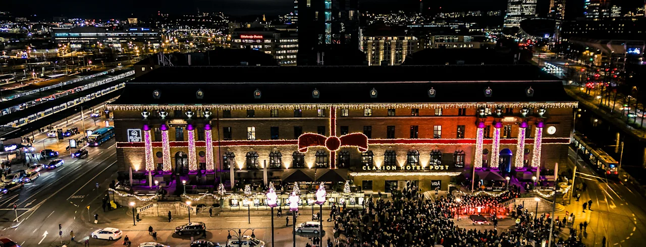 A large, ornately-lit building decorated with neon-colored lights and a giant red bow faces a bustling urban street filled with a large crowd of people gathering at nighttime.