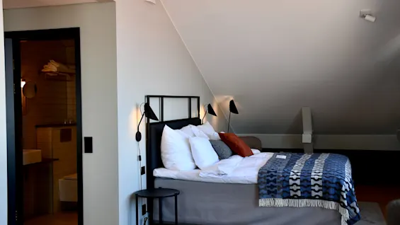 Ett mysigt hotellsovrum med en stor säng, sängbord och en blåmönstrad filt, med ett badrum synligt genom en öppen dörr.