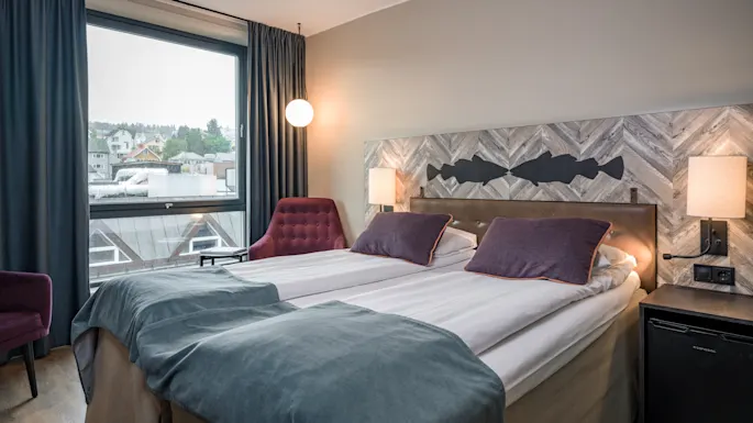 A cozy hotel room with two beds, a window view of a town, and a decorative headboard with fish silhouettes.