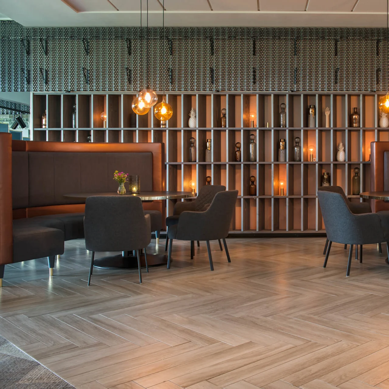 Modern dining tables with dark chairs and semicircular booths in a warm, elegant restaurant. Background features decorative shelving with ornaments, illuminated by pendant lights.
