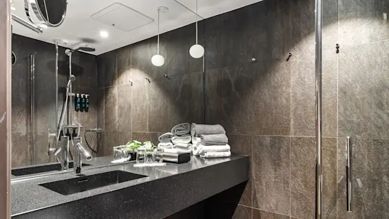 En elegant badeværelsesbordplade har pænt foldede håndklæder og glas, placeret under moderne belysning mod flisebelagte vægge. Ved siden af er et stilfuldt bruseområde med loftslamper og indbyggede shampooflasker.