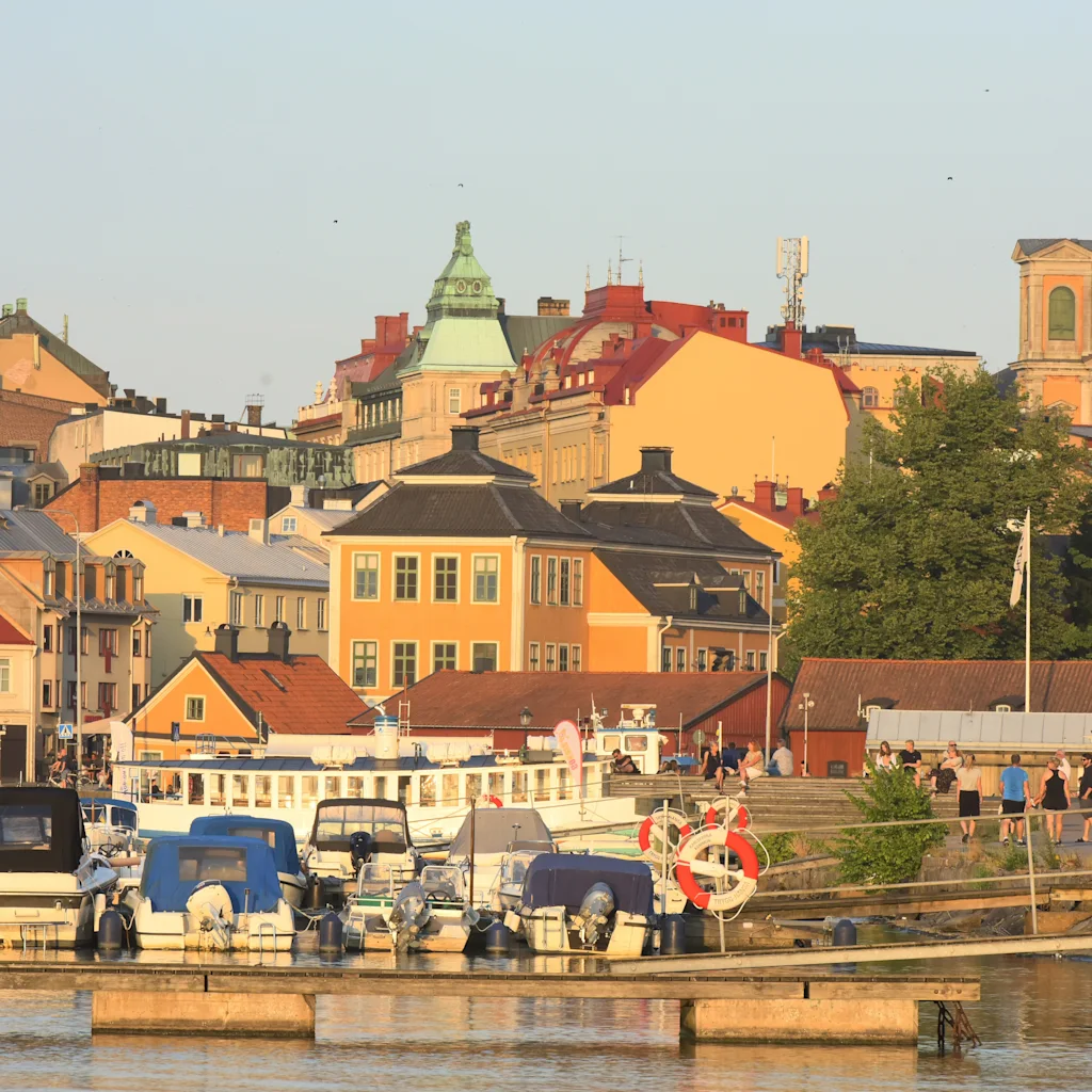A vibrant waterfront scene in Karlskrona with boats docked and people strolling along the promenade, colorful buildings in the background.