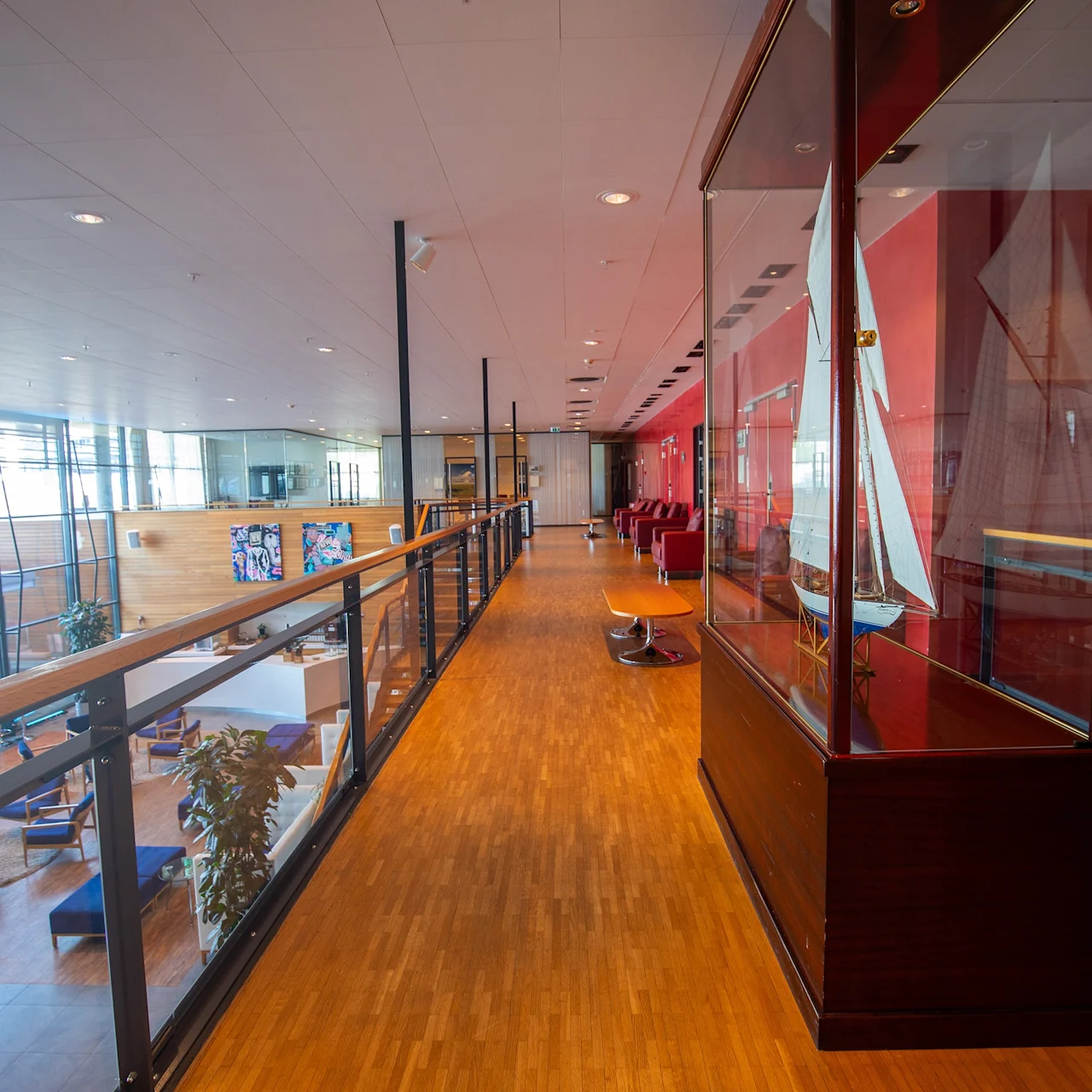En skipsmodell i en glassvitrineskap står på en gangvei med tregulv og rødmalte vegger. Gangveien har glassrekkverk og utsikt over en moderne lounge med store vinduer.