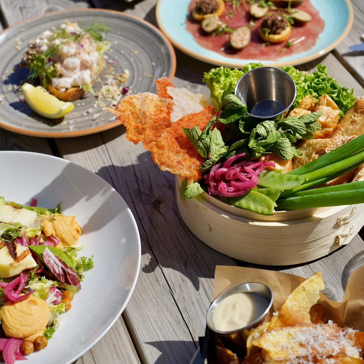 A variety of gourmet dishes arranged on a wooden table under sunlight. Plates feature cured meats, cheese, salads, and vibrant garnishes, creating an inviting outdoor dining scene.