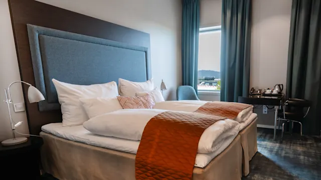Sänky, jossa on valkoiset lakanat ja oranssi peitto, on hotellihuoneessa. Lähellä on teetarjotin ja tuoli ikkunalla, josta näkyy ulkonäkymä.