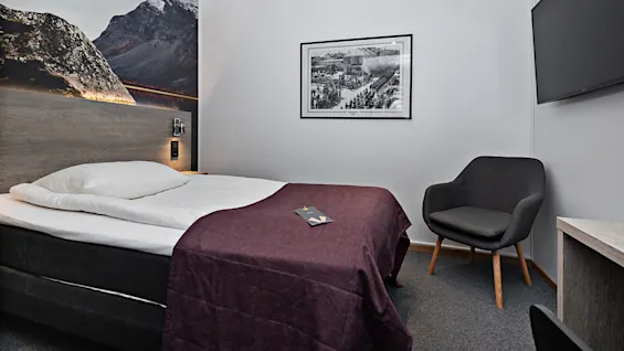 En säng med lila filt står mot en väggmålning av ett bergslandskap, bredvid en grå fåtölj och platt-TV i ett minimalistiskt rum.