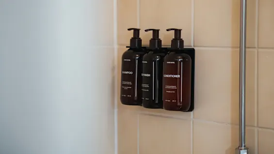 Three brown bottles labeled "HOME HOTEL SHAMPOO," "BODY WASH," and "CONDITIONER" are mounted on beige tiled bathroom wall, positioned beside a metal shower rod.
