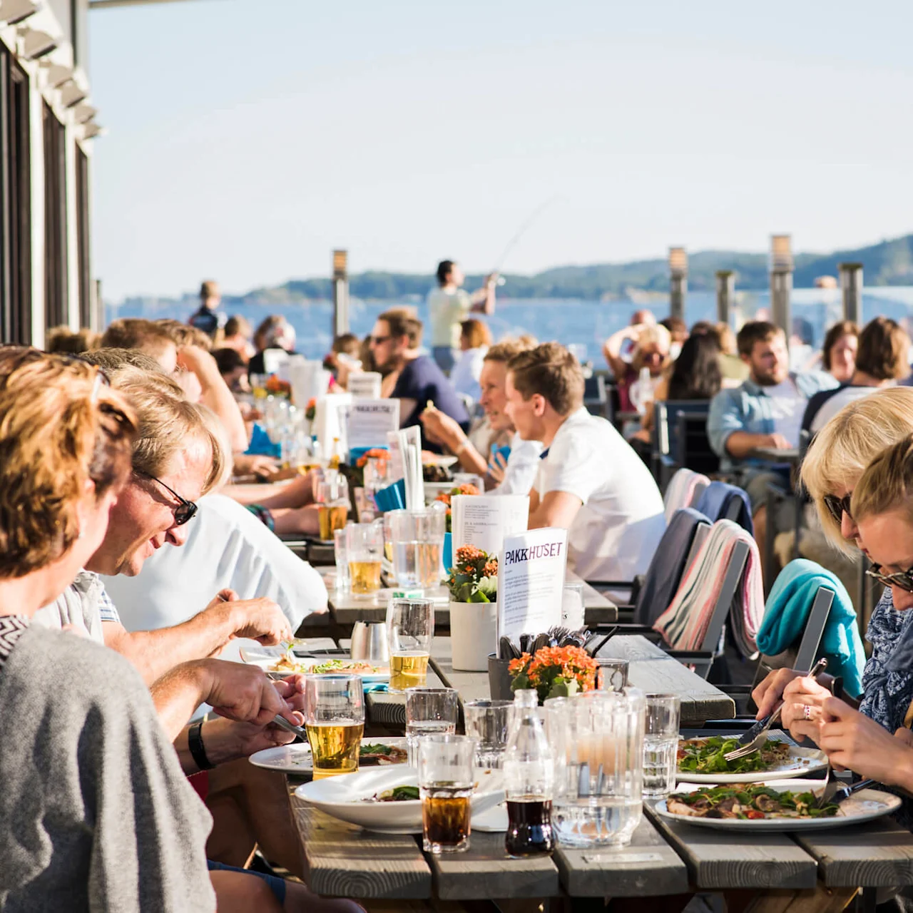 Folk spiser ved udendørs restaurantborde ved vandet på en solrig dag. Tekst: "PAKKHUSET" på bordmenuer.
