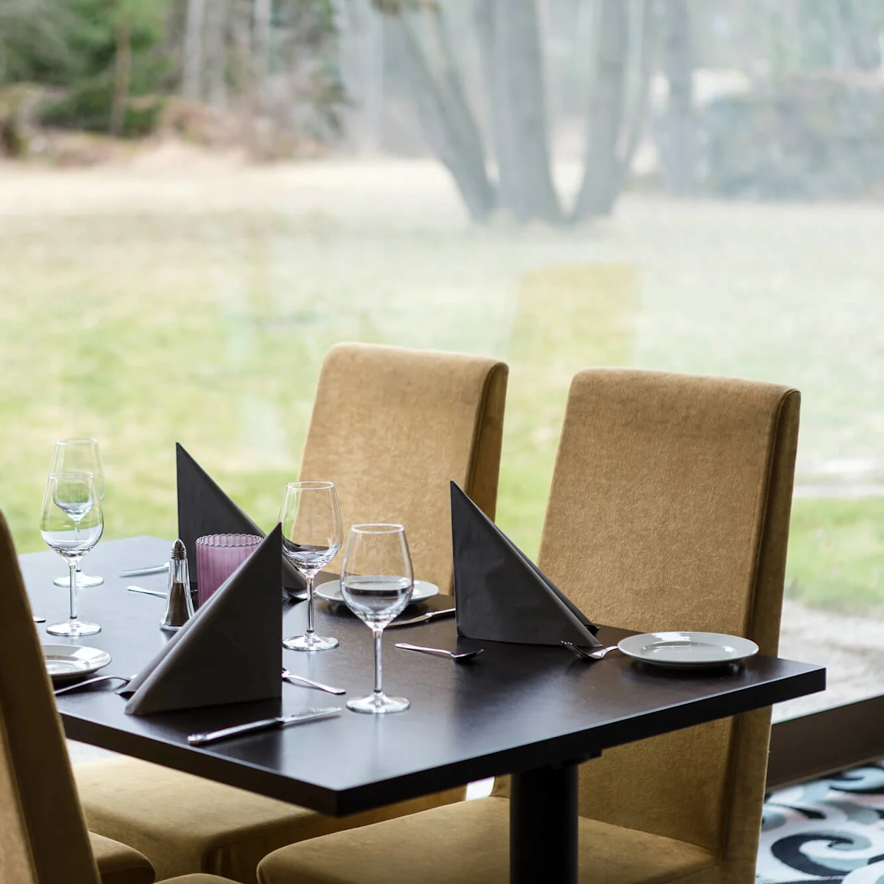 A dining table is set with wine glasses, folded napkins, and plates surrounded by beige upholstered chairs. It overlooks a grassy field through large windows.