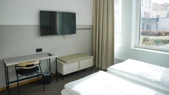 En flatskjerm-TV er montert på veggen over et skrivebord og en stol, med et vindu som viser en utsikt utenfor i et ryddig rom.