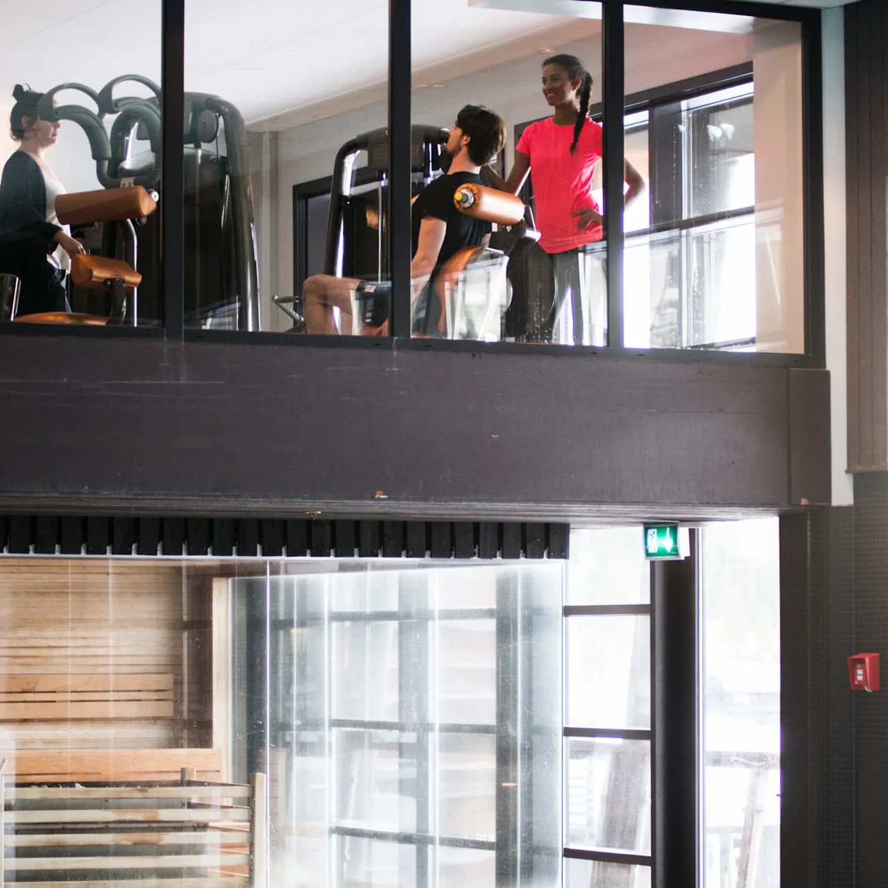 Gym-goers exercise on machines upstairs while a woman stands nearby, overlooking an adjacent swimming pool below through glass windows in a modern fitness facility.