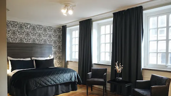 En king size-seng med svart sengetøy står mot en mønstret vegg, flankert av vinduer med mørke gardiner, to lenestoler og et lite bord med en vase i et stilrent, moderne soverom.