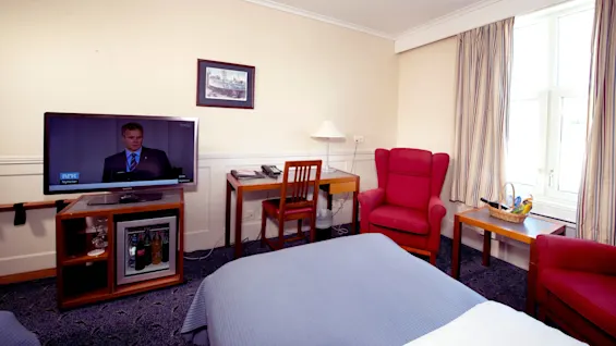 Ett hotellrum med två sängar, ett skrivbord, stolar och en TV som visar nyheter med logotypen "NRK Nyheter".