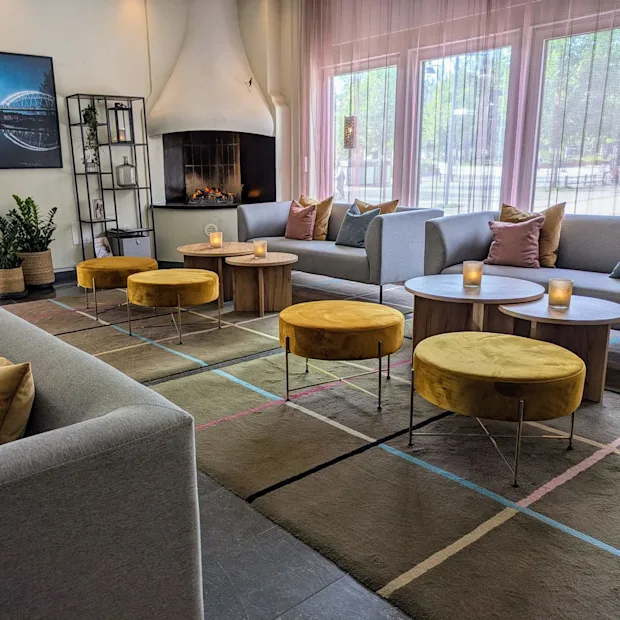 Soffor med färgglada kuddar omringar runda bord med ljus. Gula pallar står på en mönstrad matta. En öppen spis och väggkonst skapar en mysig atmosfär vid stora fönster.