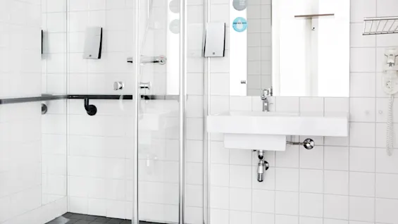 Ett vitt kaklat badrum har en glasdörr till duschen, ett handfat med spegel och en väggmonterad hårtork. Två klistermärken på spegeln lyder: ”DRINK MORE WATER” och ”GO EASY CLEAN PLANET”.