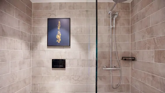 A shower with a chrome showerhead and control panel on a tiled wall; a framed abstract picture hangs next to it. The setting is a modern bathroom.