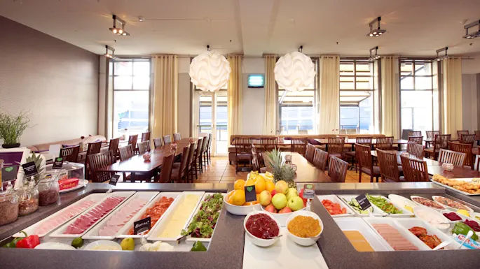 A buffet with various foods spread out, surrounded by wooden tables and chairs in a bright dining area.