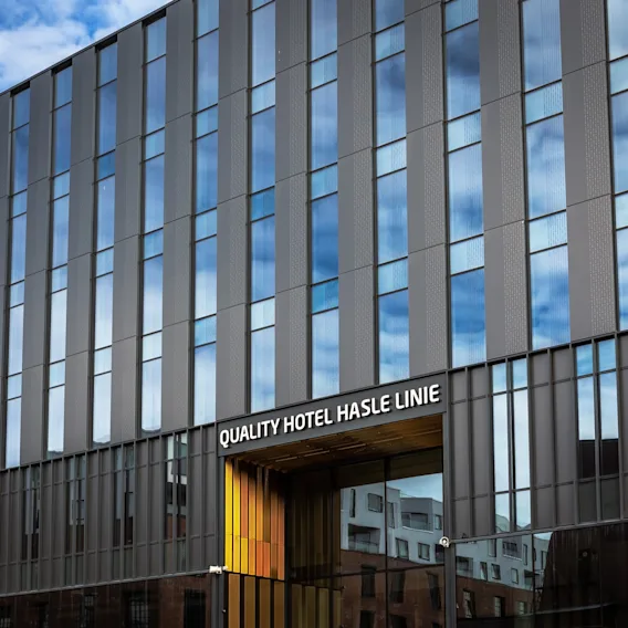 En moderne hotellfasade med reflekterende glasspaneler viser blå himmel og skyer. Teksten over inngangen lyder "QUALITY HOTEL HASLE LINIE." I nærheten er det synlig en leilighetsbygning.