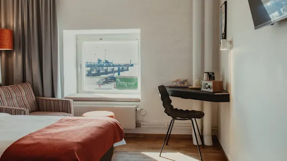 Valoisa hotellihuone, jossa on sänky, työpöytä ja ikkuna, josta on näkymä satamaan, jossa on vihreä Bring-kontti.