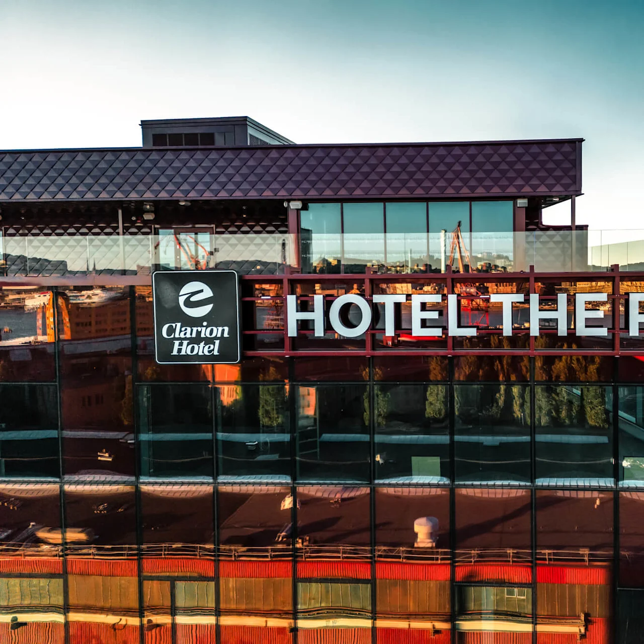 Skylten "Clarion Hotel - HOTEL THE PIER" är monterad på en glasfasad, med utsikt över en vattendrag, andra byggnader och en klar himmel i bakgrunden.