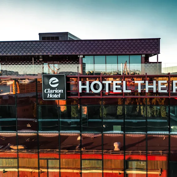 Skiltet "Clarion Hotel - HOTEL THE PIER" er montert på en glassfasade, med utsikt over en vannkant, andre strukturer og en klar himmel i bakgrunnen.
