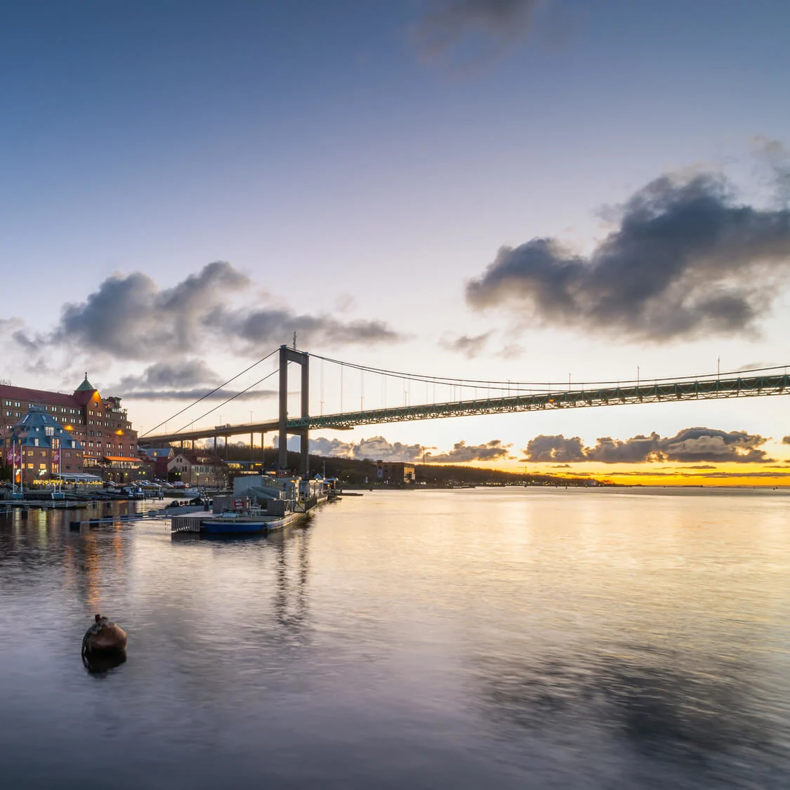 Buildings with illuminated windows line the waterfront beneath a large suspension bridge, under a cloud-dappled evening sky, reflecting on calm water where boats are docked.