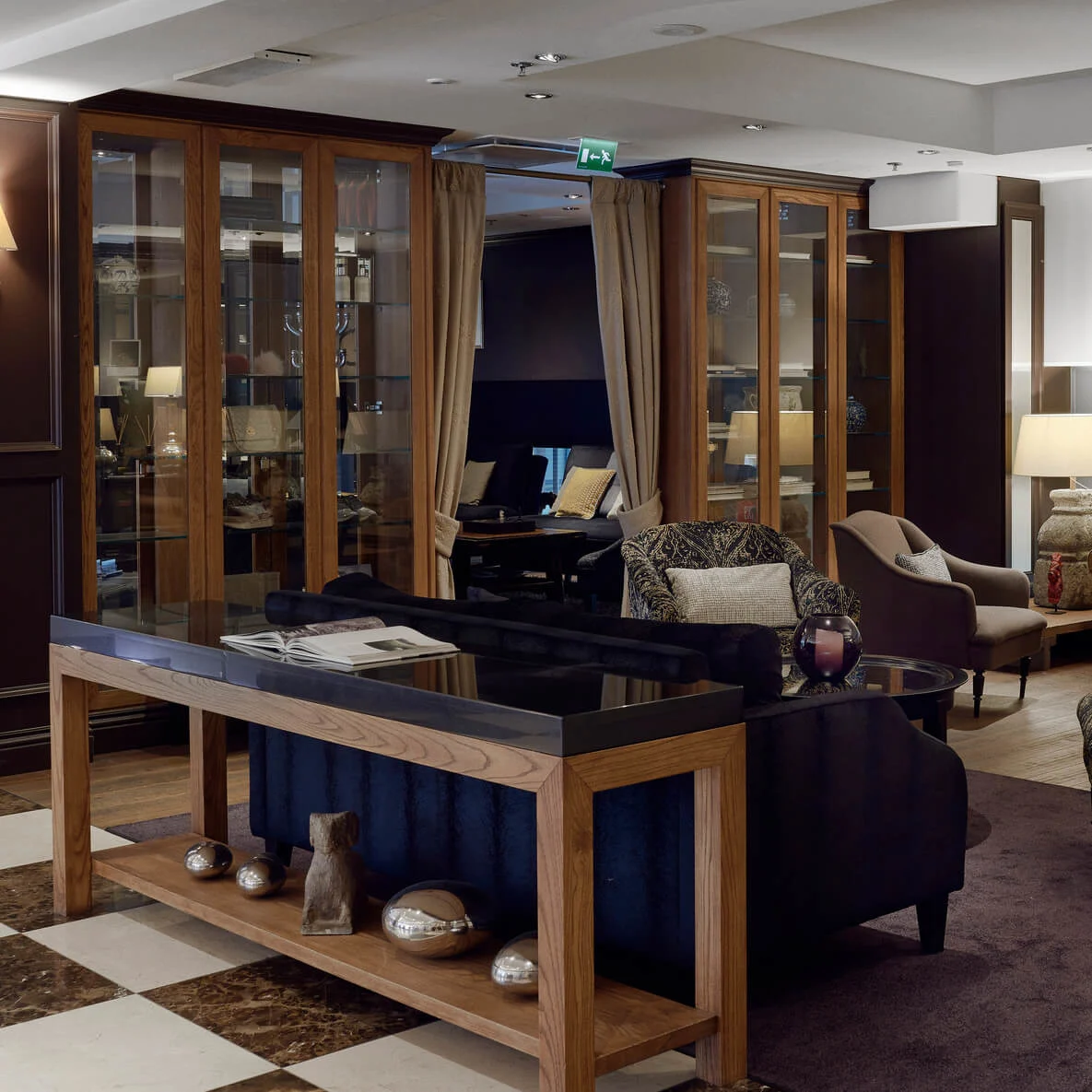 A seating area features armchairs and sofas around a coffee table. The room has dim lighting, wooden floors, and decorative items on a nearby console table and shelves.