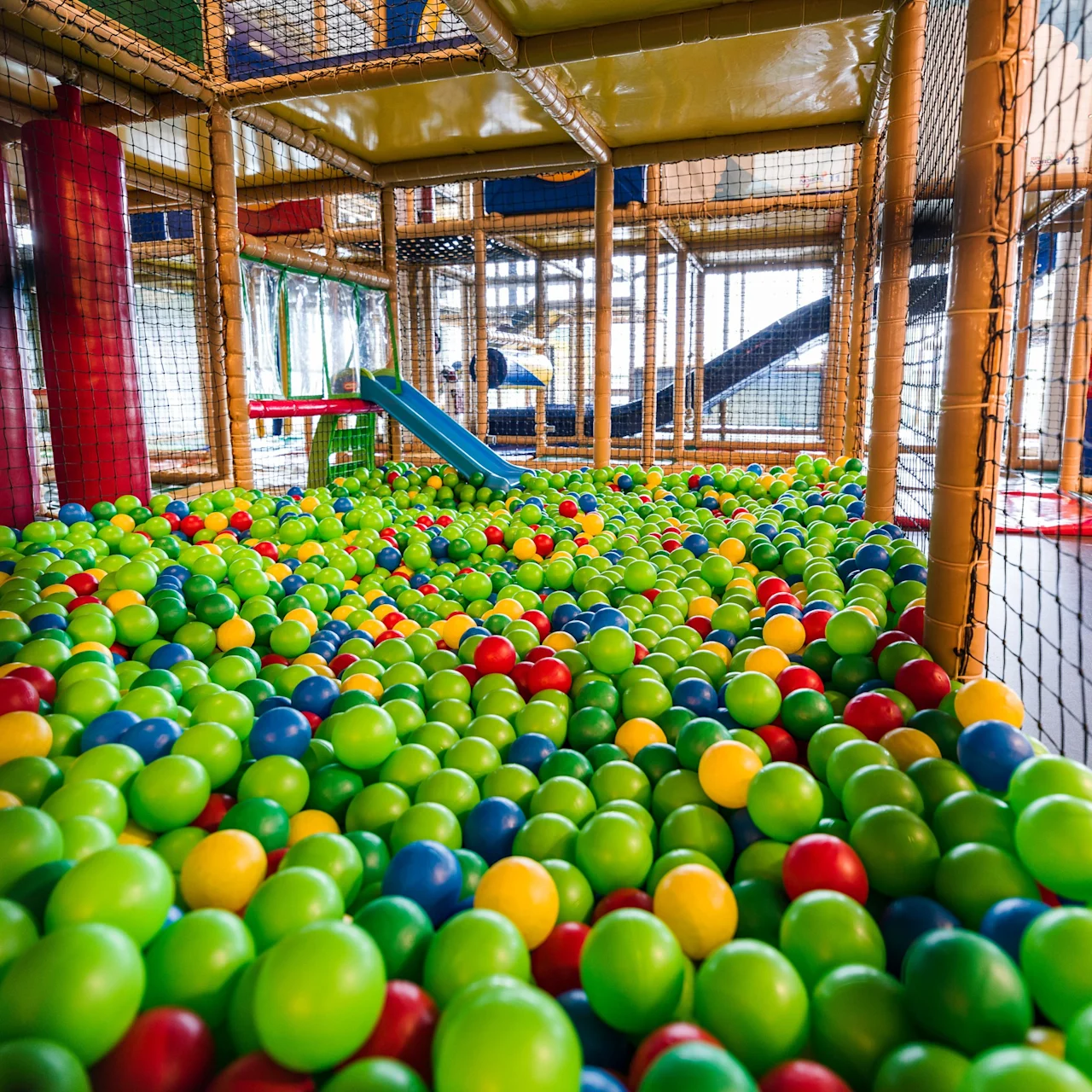 Colorful plastic balls fill a net-enclosed play area with padded structures, including a small blue slide in a recreational indoor playground setting.