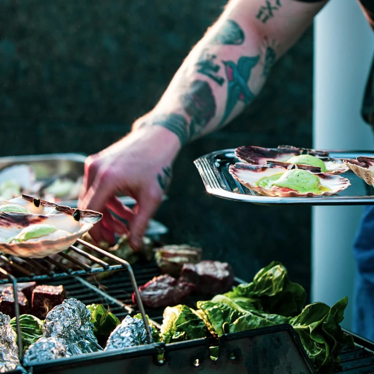 A tattooed arm grills food, placing shellfish onto a barbecue filled with leafy greens, wrapped items, and meats, in a casual outdoor cooking setting.