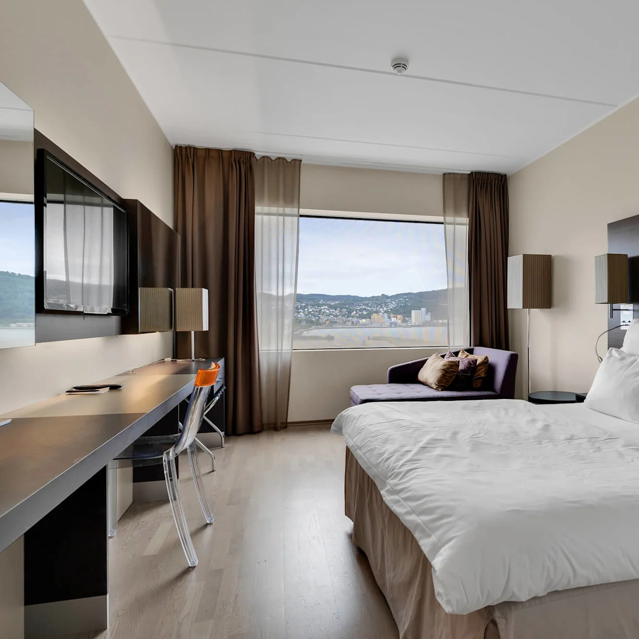 Modern hotel room features a neatly made bed with white linens beside a window overlooking a cityscape. A desk and chair are near a wall-mounted TV, surrounded by warm, neutral decor.