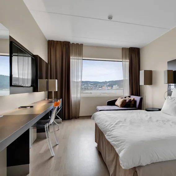 Et moderne hotellrom har en pent oppredd seng med hvitt sengetøy ved et vindu med utsikt over en by. Et skrivebord og stol er ved en veggmontert TV, omgitt av varme, nøytrale dekorasjoner.