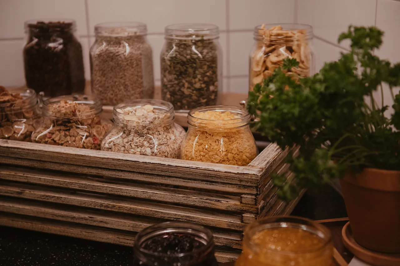 A cozy breakfast spread featuring various cereals, grains, and dried fruits in glass jars, neatly arranged in a rustic wooden tray.