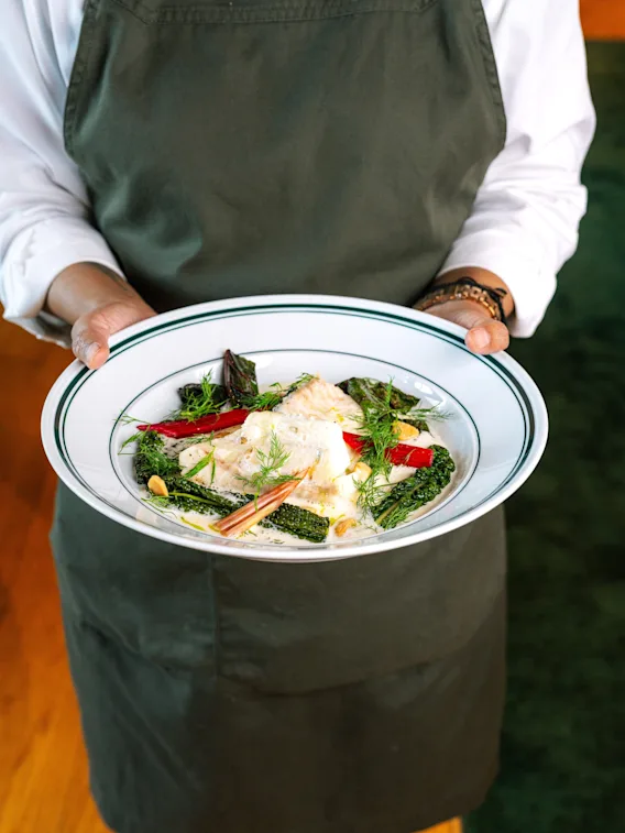 A person in a green apron holding a white plate with a delicious-looking meal of fish, green vegetables, and red peppers.