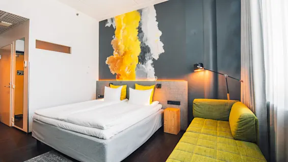 Bed resting beside a yellow, gray, and white abstract wall mural, with green sofa and floor lamp in a hotel room. Light enters through a large window with sheer curtains.