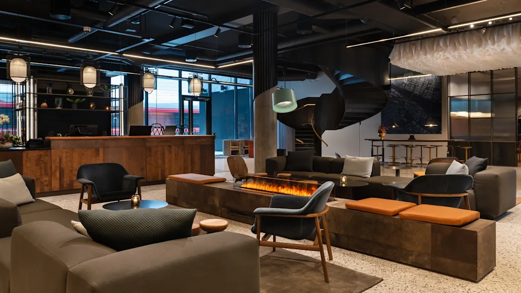 A modern lobby with stylish seating surrounds a sleek, rectangular fireplace. Overhead, industrial lights hang from the ceiling. A wooden reception desk and spiral staircase are visible.